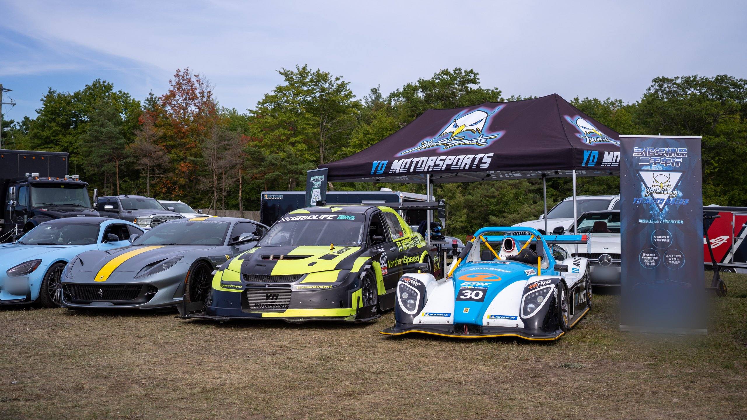 YD Motorsports exhibitor stand at the Drive Festival, Canadian Tire Motorsport Park, Bowmanville, Ontario, in September 2022: a Mercedes-AMG GT, a Ferrari 812 Superfast, the YD Motorsports Mitsubishi Lancer Evolution and a Radical race car in front of the team tent and a YD Auto Sales banner.