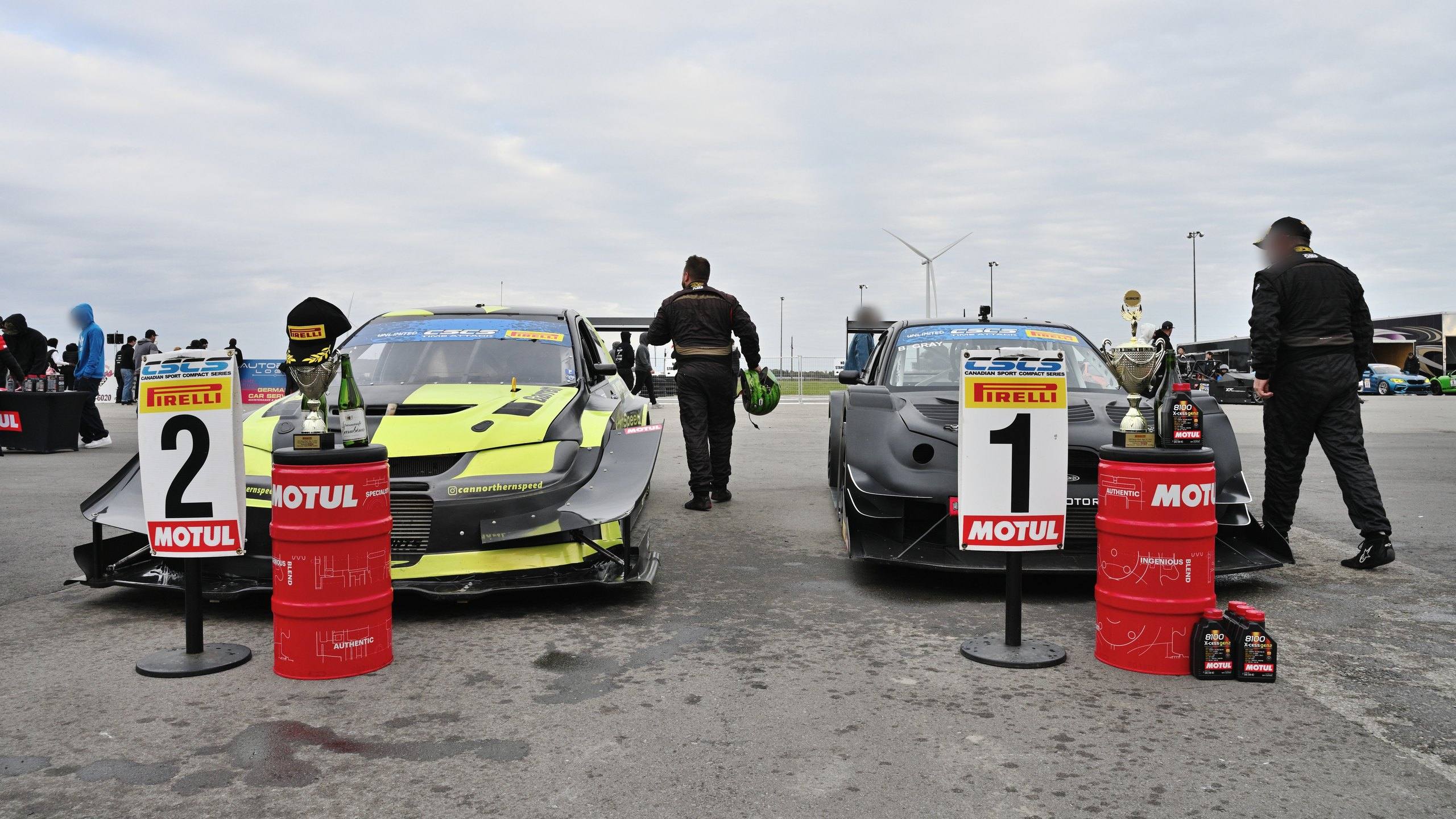 Podium line-up at the Canadian Sport Compact Series time attack finale in October 2022: the YD Motorsports-liveried Mitsubishi Lancer Evolution beside the second-place board and the winning car beside the first-place board, with trophies set on Motul oil drums.