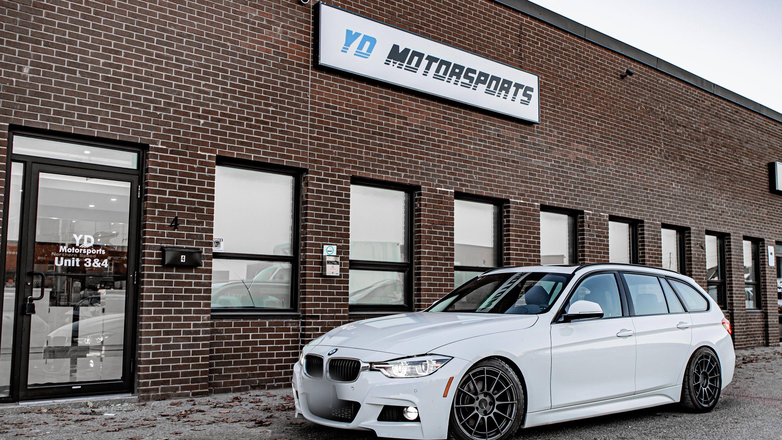 The first YD Motorsports workshop in Vaughan, Ontario, in November 2020: a brick industrial building with the YD Motorsports sign above the windows and a door decal reading Unit 3&4, with a white BMW 3 Series Touring parked in front.