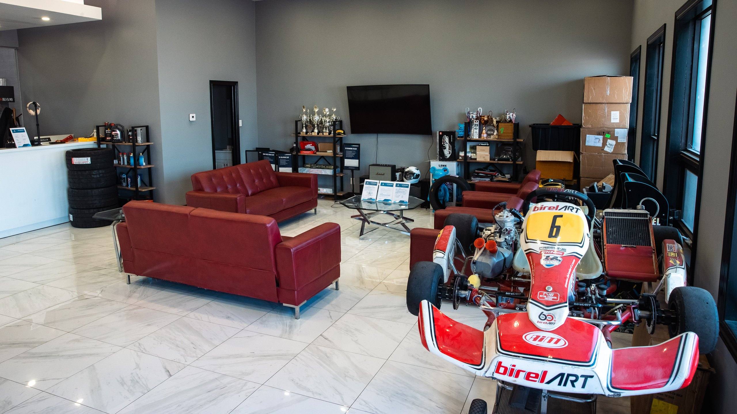 Customer lounge at the YD Motorsports workshop in September 2023: red leather sofas, shelves of racing trophies, a wall-mounted screen and a Birel ART racing kart on a white tiled floor.