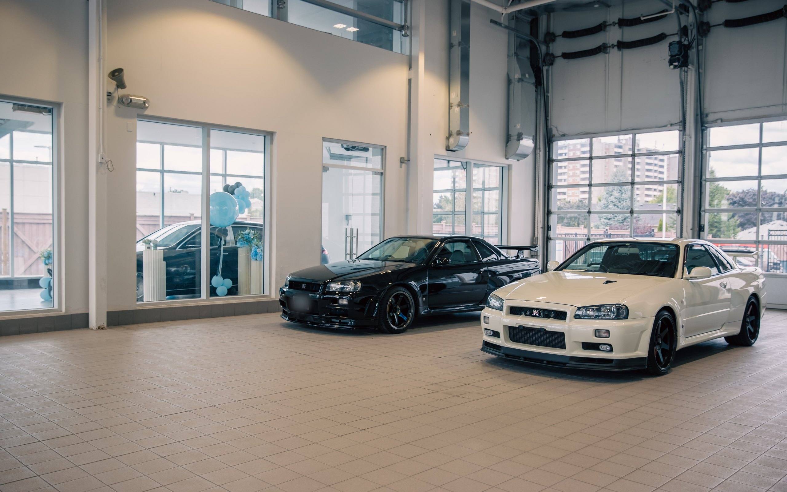 Two Nissan Skyline GT-R (R34) coupés, one black and one white, in a tiled bay with glazed roll-up doors at the Newmarket headquarters.