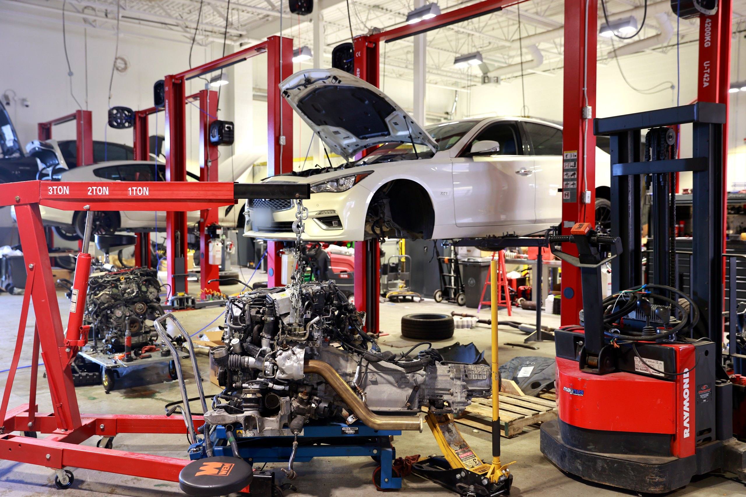 Mechanical workshop with a removed engine and transmission assembly on a support stand, an engine hoist, and an Infiniti Q50 raised on a lift behind.
