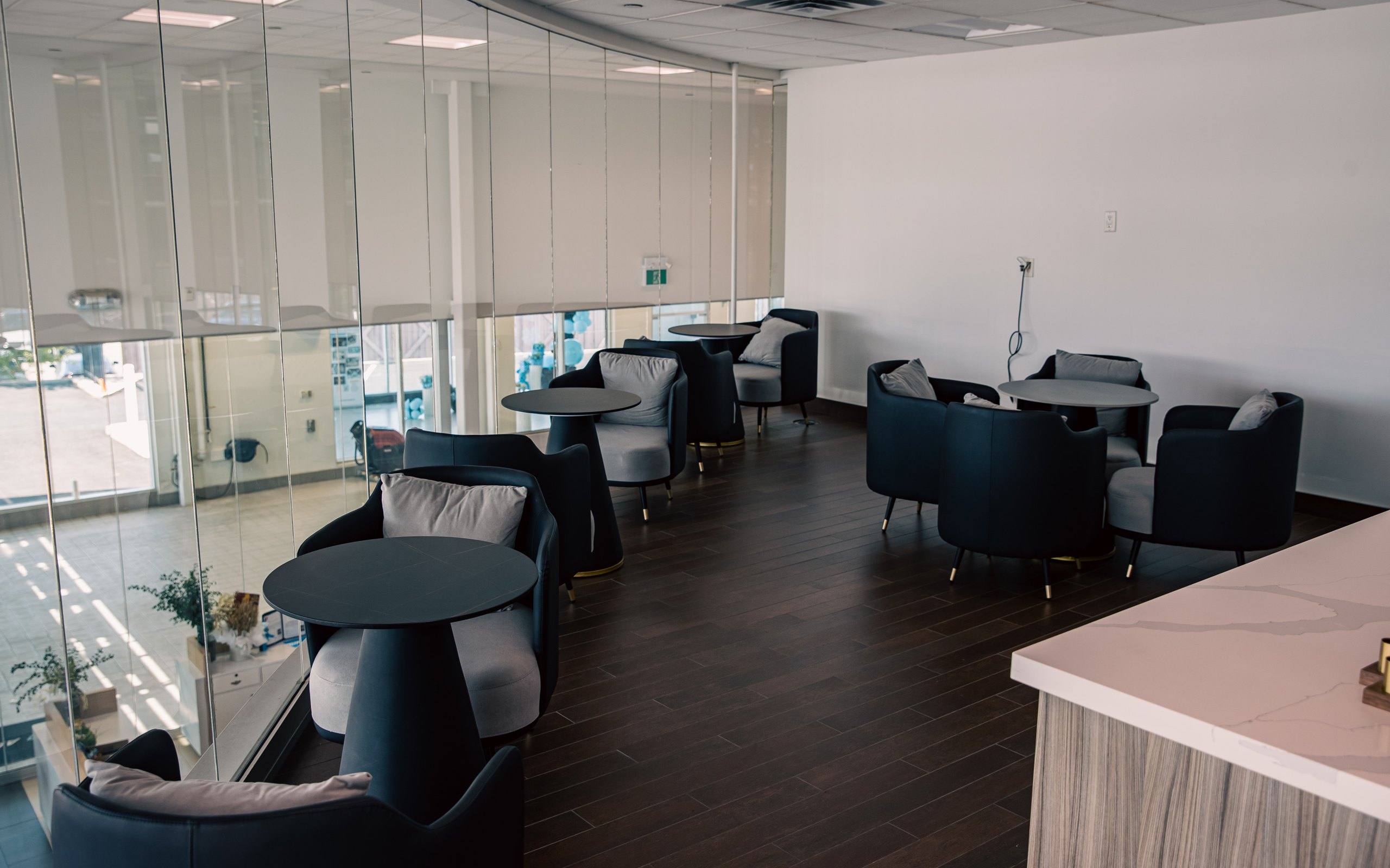 Customer lounge on the mezzanine with upholstered armchairs and round tables, overlooking the ground floor through a curved glass wall.
