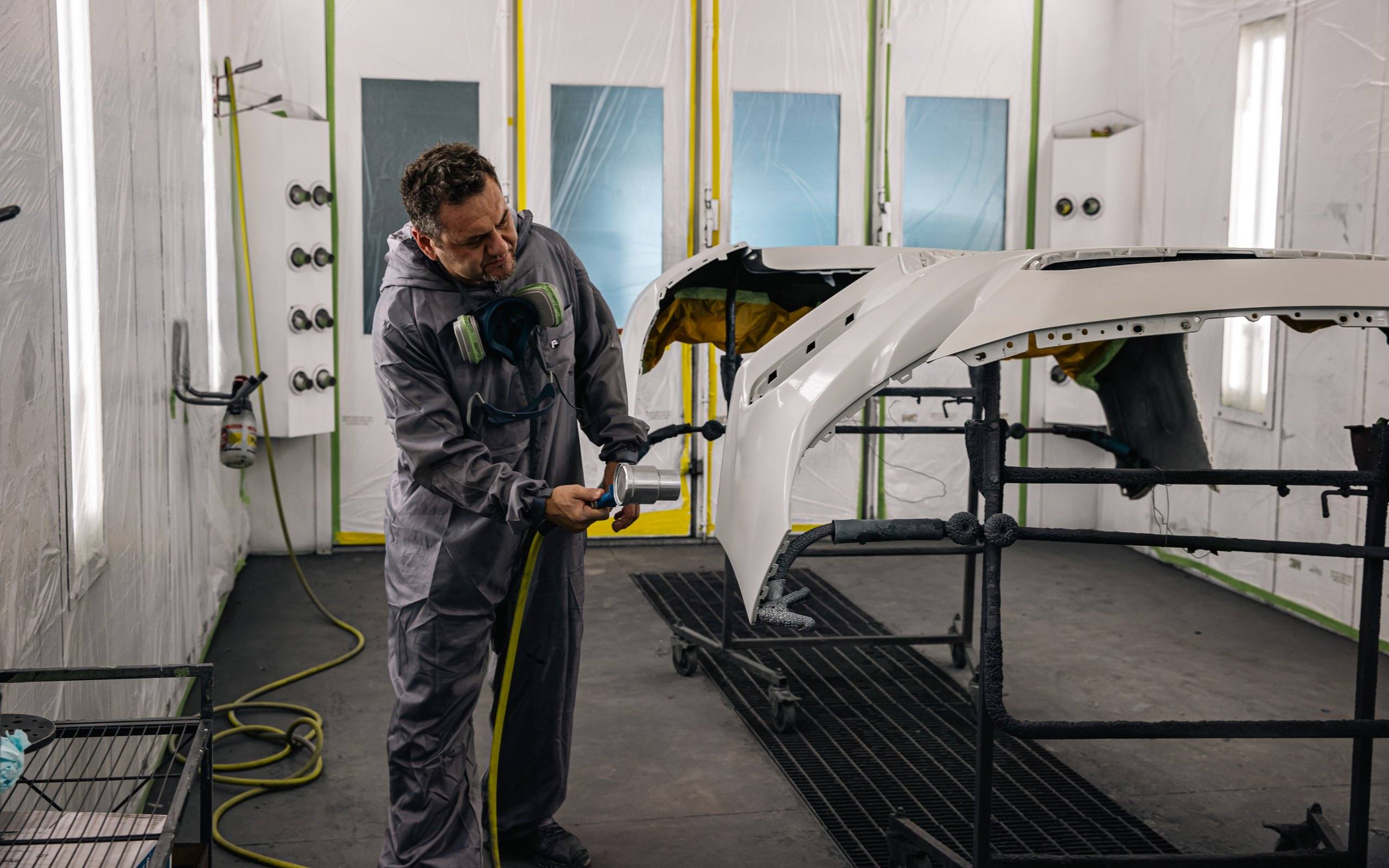 A painter in protective coveralls with a respirator works with a spray gun on freshly painted white bumper covers inside the paint booth.