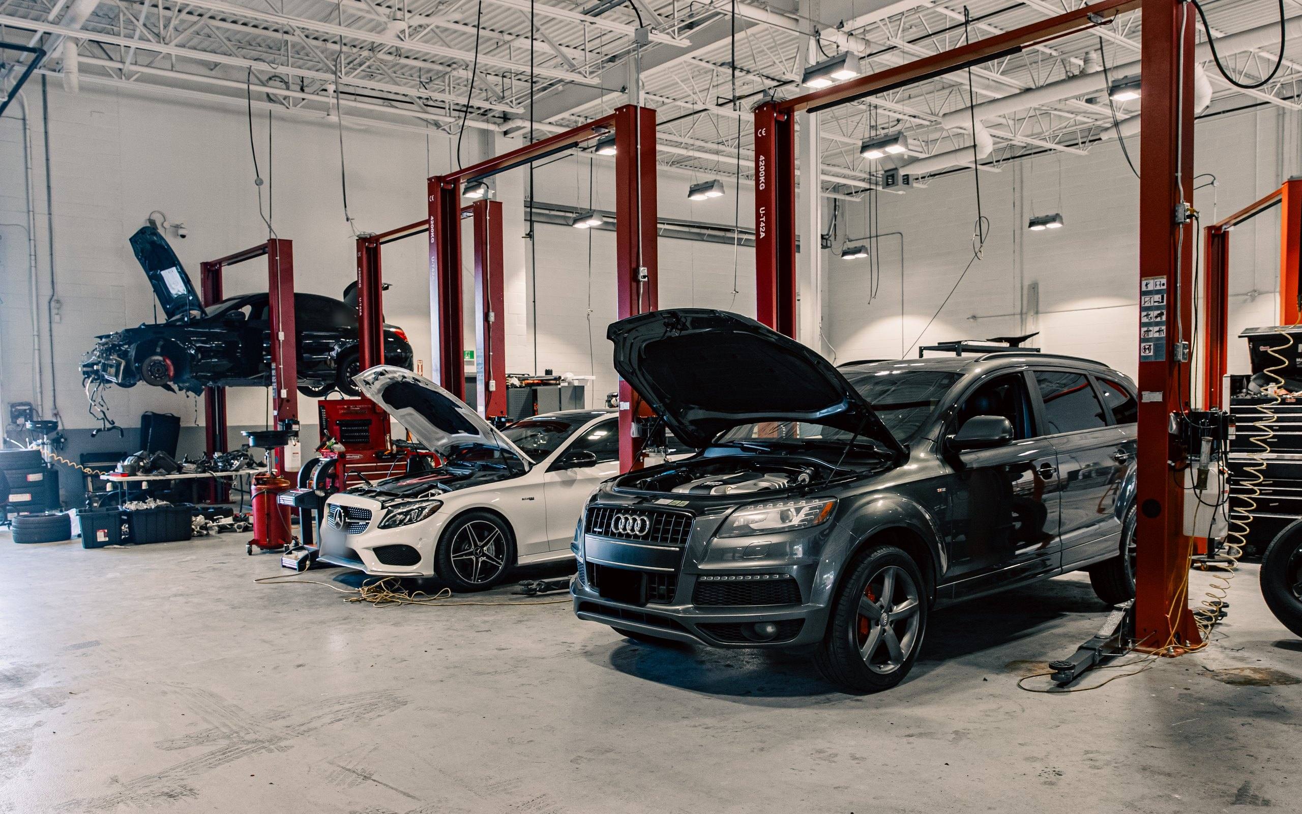 Service bays with an Audi Q7 and a Mercedes-Benz C-Class under inspection with bonnets open, and a collision-damaged sedan raised on a two-post lift behind.
