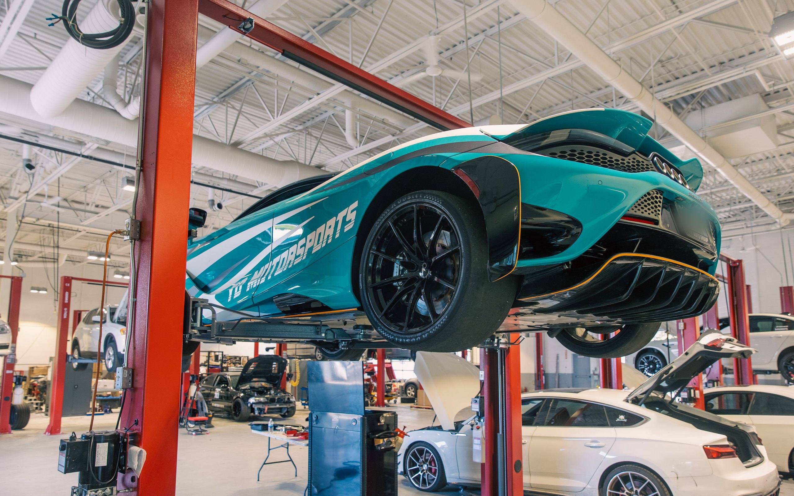 Service workshop with a teal McLaren in YD Motorsports livery raised on a two-post lift and further vehicles in the service bays behind.