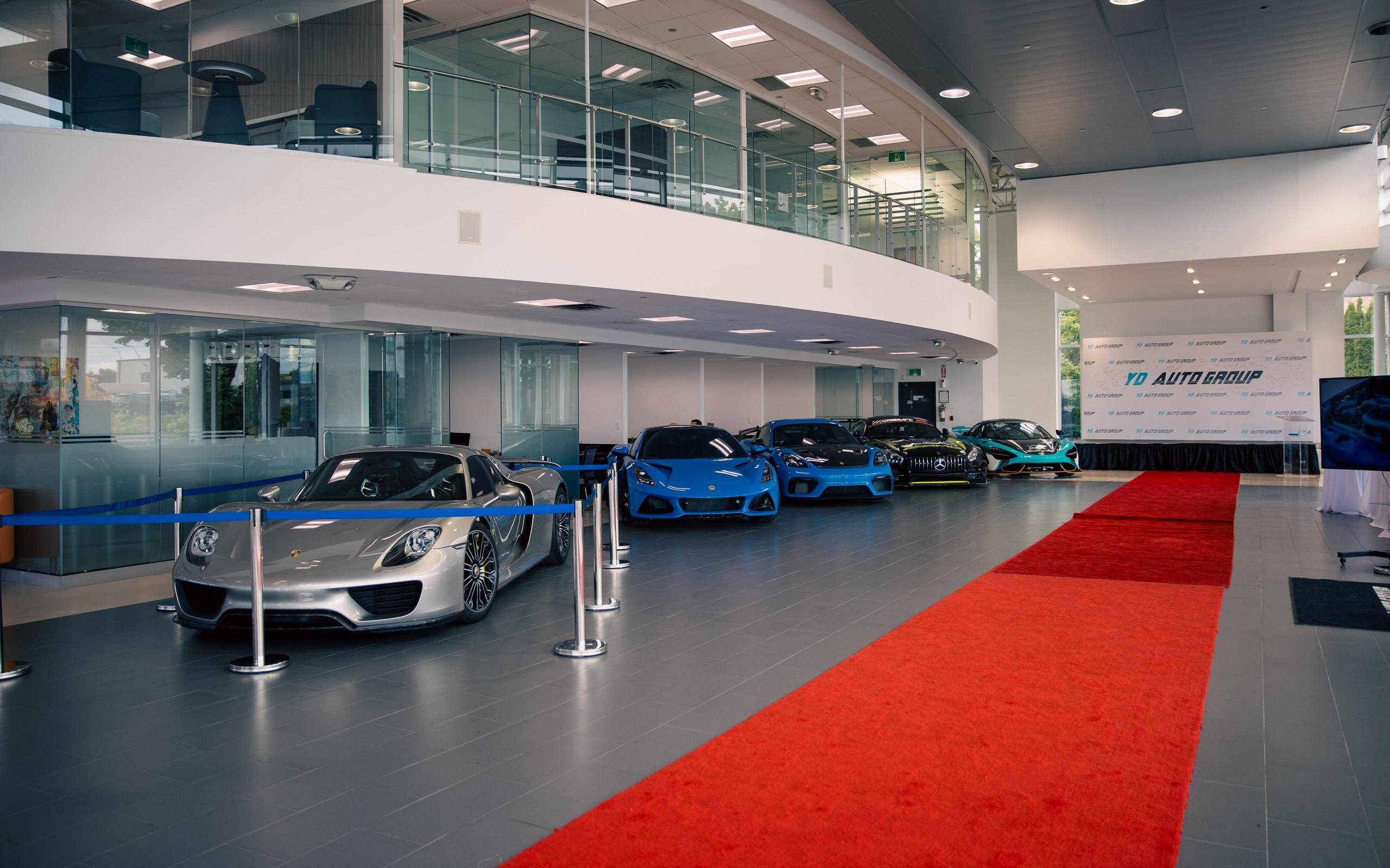Double-height showroom at the YD Auto Group headquarters: a Porsche 918 Spyder leads a line of performance cars including a Lotus Emira, a Porsche Cayman, a Mercedes-AMG GT and a McLaren beside a red carpet, below the glass-fronted mezzanine offices.