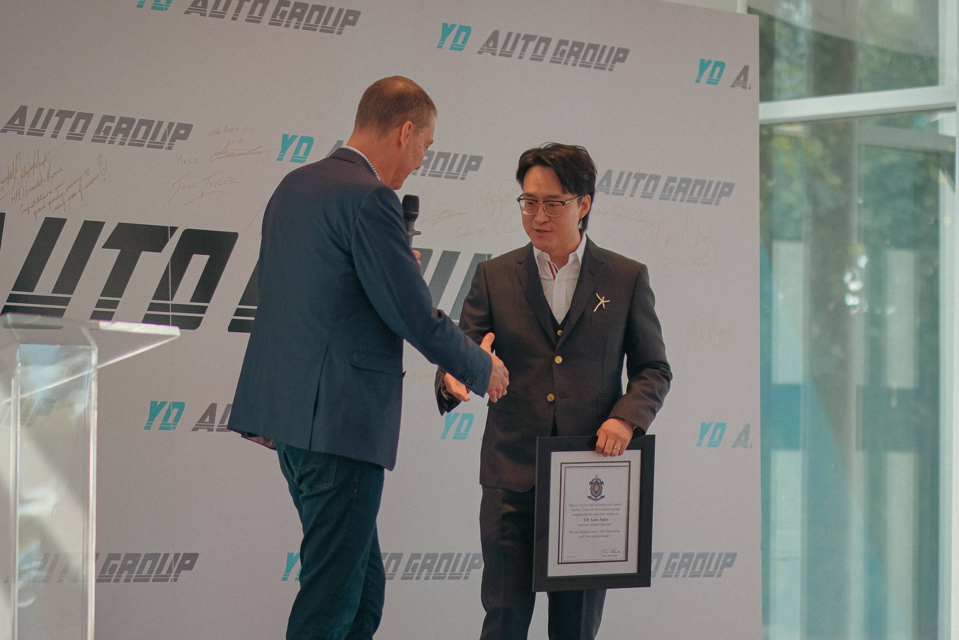 On stage at the July 18, 2024 grand opening, YD Auto Group receives a framed certificate of congratulations from Mayor John Taylor and Members of Council of the Town of Newmarket.